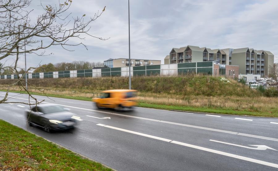 Trafikeret vej i forgrunden for boliger, der er omkranset af en støjmur